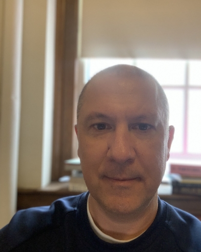 A close-up of a man with a shaved head wearing a dark shirt, sitting indoors near a window with soft natural light filtering through blinds. The background includes a bookshelf and soft focus details of a wooden frame.